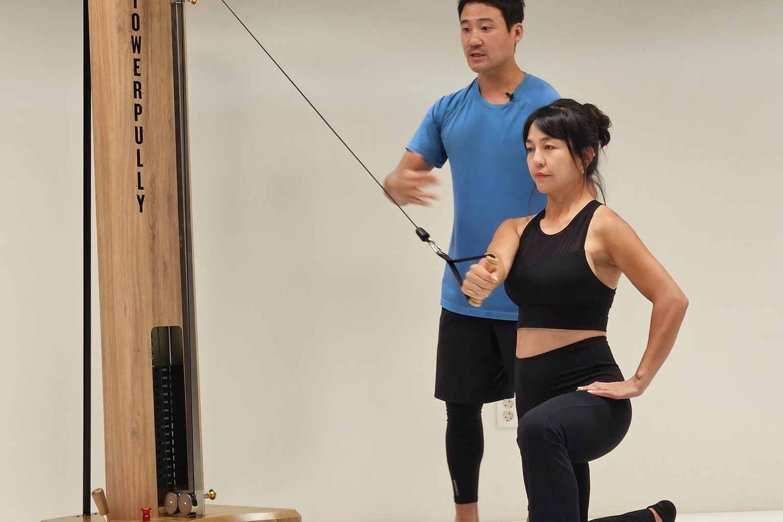 한쪽 무릎을 꿇고 옆에서 당기는 로프에 맞서 손잡이를 가슴 앞에 유지하는 동작과 이를 지도하는 강사
